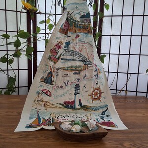 Peut inclure: Une nappe sur le thème de Cape Cod de couleur crème avec un motif de carte représentant un phare, un pont et un voilier. La nappe a un bol décoratif en bois avec des coquillages au premier plan. Le texte "Cape Cod" est visible.