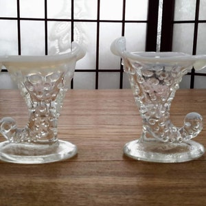 Peut inclure: Deux vases en verre transparent en forme de corne d'abondance avec une surface texturée et bullée. Les vases ont une finition opalescente blanche et sont posés sur une surface en bois.