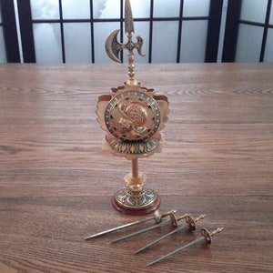 May include: A gold-toned metal decorative stand with a shield design and a spear with a crescent-shaped blade. The stand has a round base and three legs. Four silver-toned metal skewers are lying on a wooden surface.