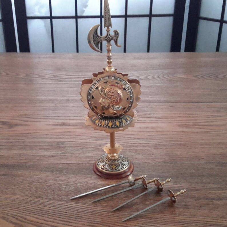 May include: A gold-toned metal decorative stand with a shield design and a spear with a crescent-shaped blade. The stand has a round base and three legs. Four silver-toned metal skewers are lying on a wooden surface.