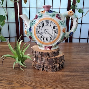 May include: A decorative ceramic teapot with a clock face design. The teapot is white with floral and berry accents, and a pink lid. It sits on a stack of wooden slices. The clock face has black numbers and hands.