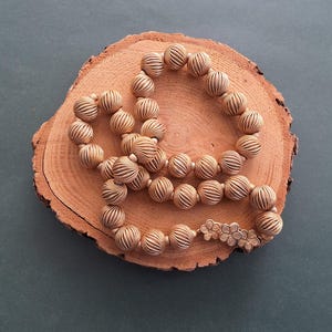 May include: A gold-colored necklace made of woven fabric beads. The beads are round and have a textured surface. The necklace is displayed on a wooden surface.