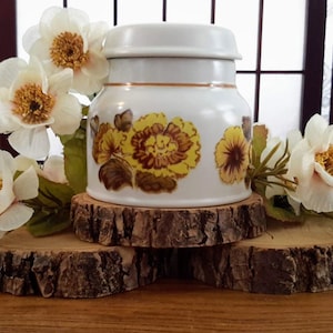 Peut inclure: Un pot en céramique blanche avec un motif floral composé de fleurs jaunes et brunes. Le pot est posé sur trois tranches de bois, et des fleurs blanches sont disposées autour du pot.