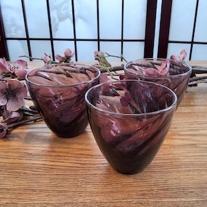 May include: Three purple and brown swirl-patterned glass tumblers. The glasses are set on a wooden surface, with floral accents in the background. The glasses have a unique, swirled design.