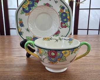 May include: A vintage teacup and saucer set. The cup and saucer are decorated with colorful floral patterns in blue, yellow, and red. The cup has a green handle and a white base. The saucer has a unique shape with a green border.