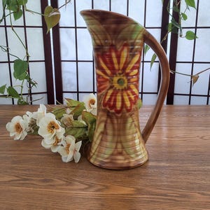 May include: A decorative pitcher with a brown handle and a flared base. The pitcher has a large, stylized flower design in red, yellow, and green. The background is a wooden surface with white flowers and foliage.