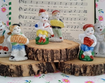 May include: A collection of seven small porcelain figurines of children, each dressed in colorful outfits and holding musical instruments or props. The figurines are arranged on a wooden display, with sheet music in the background.