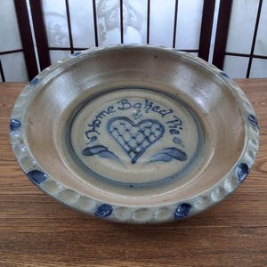 May include: A round, tan ceramic pie dish with a scalloped edge featuring blue painted accents. The centre of the dish displays a blue heart graphic with a cross-hatched pattern and the text Home Baked Pie written in a cursive script font.