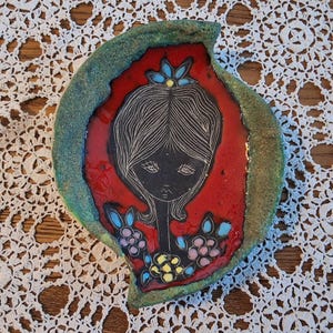 May include: A ceramic dish with a red centre featuring a black silhouette of a woman's face and floral accents. The dish has a green and blue textured border. The design is reminiscent of vintage art.