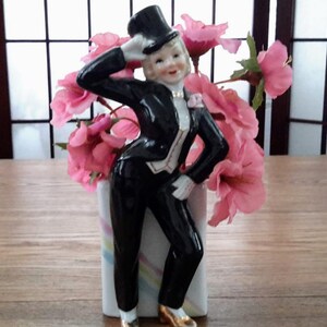 Peut inclure: Une figurine en porcelaine noire et blanche d'une personne portant un smoking et un chapeau haut de forme. La figurine tient une fleur et se tient dans un vase blanc avec des fleurs roses.