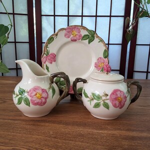 May include: A cream-coloured ceramic tea set featuring pink rose floral designs. The set includes a creamer, a sugar bowl with a lid, and a plate. The handles and rims have a brown, textured finish. The floral pattern includes green leaves.