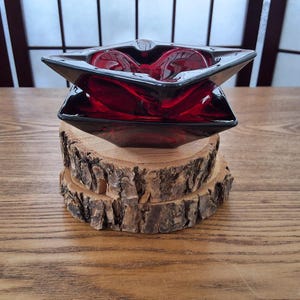 Peut inclure: Un cendrier en verre rouge foncé avec un design angulaire unique. Le cendrier est posé sur une base en bois.