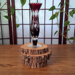 May include: A tall, ruby red glass vase with a clear, etched floral design. The vase has a flared top and a clear, circular base. It is displayed on a rustic, wooden slice stand. The vase is approximately 10 inches tall.