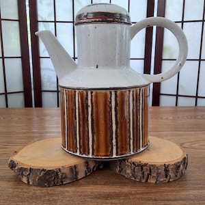 Peut inclure: Une théière en céramique avec une partie supérieure blanc cassé mouchetée et une base cylindrique à rayures verticales brunes et blanches. L'objet comprend un couvercle assorti, une anse incurvée et un bec verseur pointu, le tout vernissé.