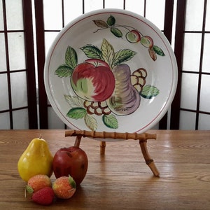 Peut inclure: Une assiette en céramique blanche avec un motif peint à la main de fruits, y compris des pommes, des poires et des raisins. L'assiette est sur un support en bambou et entourée de fruits.