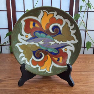 May include: A decorative ceramic plate with a colorful floral design. The plate features a green border and a central pattern with orange, yellow, blue, purple, and brown elements. It is displayed on a black stand, showcasing its intricate details.