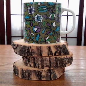 May include: A set of four wooden coasters made from tree bark, stacked on top of each other. A vintage mug with a floral pattern is sitting on top of the coasters.
