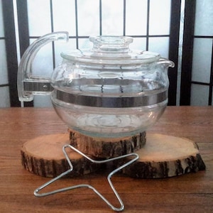 Peut inclure: Une théière en verre transparent avec une bande métallique autour du milieu, posée sur un support en bois avec un chauffe-plat en métal en dessous.