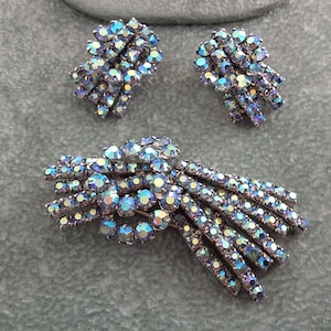 Peut inclure: Un ensemble de bijoux composé d'une broche et d'une paire de boucles d'oreilles assorties. Chaque pièce est ornée de strass irisés multicolores disposés en grappes et en éventail, reflétant des tons bleus, violets et dorés.