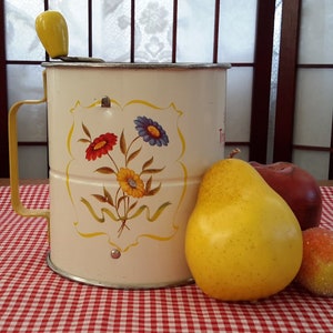 Peut inclure: Un tamis à farine vintage en métal blanc avec une poignée jaune et un motif floral. Le tamis est posé sur une nappe à carreaux rouge et blanc avec une poire jaune et une pomme rouge à côté.