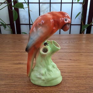 May include: A ceramic parrot ornament in orange, red, and green. The parrot has a detailed beak and eye, perched on a green base with a hollow opening. The figurine is a decorative item.