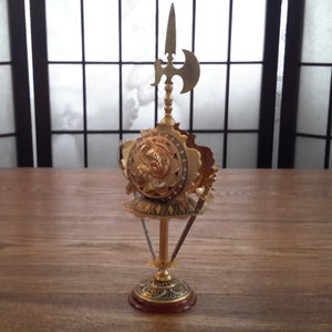 May include: A gold-colored metal decorative stand with a round top and three legs. The top features a shield with a face and a spear with an axe on top.
