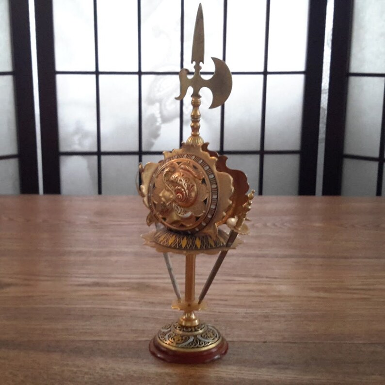 May include: A gold-colored metal decorative stand with a round top and three legs. The top features a shield with a face and a spear with an axe on top.