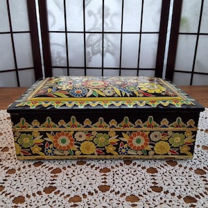 May include: A rectangular black metal box with a floral design. The lid and sides are adorned with colorful flowers and gold trim. The box sits on a white lace tablecloth.