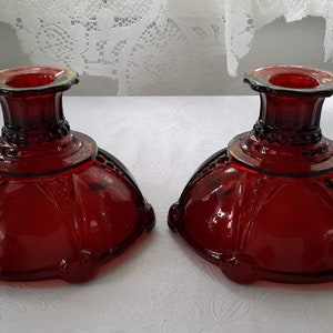 Peut inclure: Deux bougeoirs en verre rouge avec un design décoratif. Les bougeoirs ont une base large et un sommet étroit avec un bord surélevé.