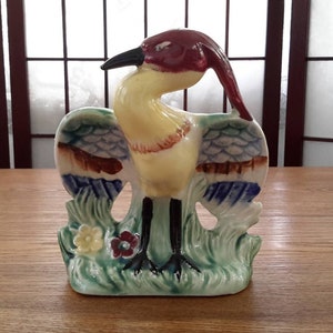 May include: A ceramic figurine of a yellow bird with red, blue and green accents. The bird is standing on a green base with flowers.