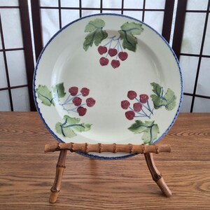 Peut inclure: Une assiette en céramique blanche avec un bord bleu et un motif de cerises peint à la main. L'assiette est soutenue par un présentoir en bambou. Les cerises sont rouges et les feuilles vertes.