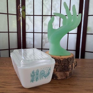 Peut inclure: Un plat de réfrigérateur rectangulaire en verre opalin blanc avec un couvercle en verre transparent nervuré. Le côté du plat présente un motif graphique sarcelle représentant un coq entouré d'épis de blé et de petites étoiles. Un présentoir en forme de main vert clair mat.