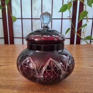 Peut inclure: Un pot en verre rouge rubis avec un couvercle assorti et un bouton en verre transparent. Le pot présente des motifs complexes en verre taillé. Le pot est posé sur une surface en bois. C'est un objet décoratif.