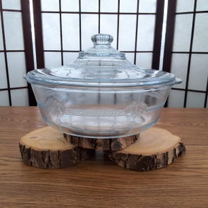 Peut inclure: Un plat à four en verre transparent avec un couvercle, orné d'un motif complexe. Le plat est posé sur deux rondins de bois.