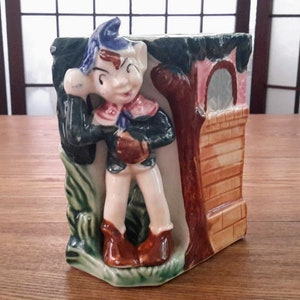 Peut inclure: Un pot en céramique en forme de petit lutin debout à côté d'un bâtiment en briques avec une porte rose. Le lutin porte un chapeau bleu, une veste verte et des bottes brunes. Le pot est décoré de feuilles vertes et brunes.