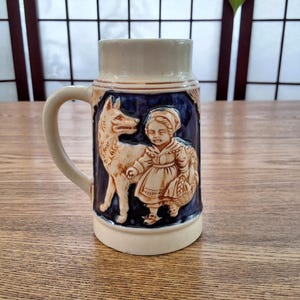 Peut inclure: Une chope à bière en céramique avec une poignée, présentant un motif en relief du Petit Chaperon rouge et du loup. La chope a un dessus et une poignée de couleur crème, avec un fond bleu pour le relief. La base est également de couleur crème.