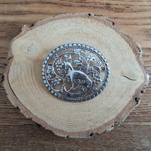 Peut inclure: Broche ovale argentée représentant une chèvre dans un motif de vigne. La broche a une bordure ajourée détaillée et est posée sur une tranche de bois naturelle. Un accessoire vintage.