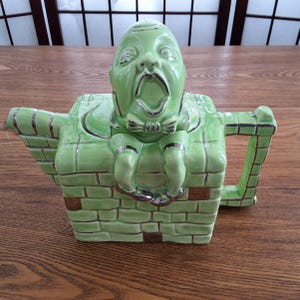 May include: A green ceramic teapot shaped like a brick chimney with a green man figure sitting on top. The man has a bow tie and is wearing silver shoes. The teapot has silver accents.