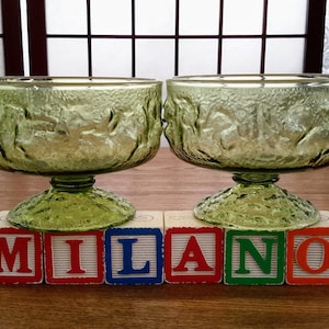 May include: Two green glass bowls with a textured design and a pedestal base. The bowls are sitting on top of wooden blocks that spell out the word "MILANO".