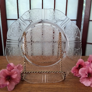 Peut inclure: Une assiette en verre transparent avec un motif floral délicat. L'assiette est sur un support en métal doré et entourée de fleurs roses.