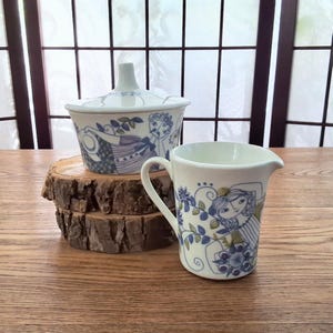 May include: A white ceramic creamer and a lidded sugar bowl featuring blue and green illustrations of stylized figures and floral patterns. The creamer has a handle and a spout, while the sugar bowl has a matching lid with a central knob.