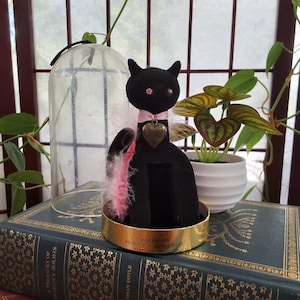 Peut inclure: Figurine de chat en velours noir avec des détails roses, un pendentif cœur doré et une plume rose. Le chat est dans une base dorée avec le texte "MAX FACTOR PRIMITIF SOPHISTI-CAT PERFUME 1931".