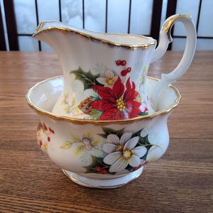 Peut inclure: Un ensemble de crémier et de sucrier en porcelaine blanche avec une bordure dorée et un motif floral représentant des poinsettias, du houx et des baies.