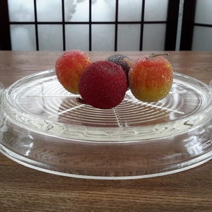 May include: A clear glass serving dish with two handles. The dish has a raised centre section with a circular pattern. Three red and yellow fruit are on the dish.