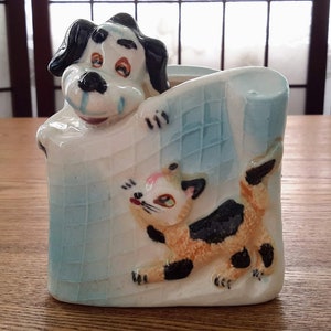 Peut inclure: Un pot de fleurs en céramique avec un motif de chien et de chat. Le chien, avec des oreilles et des taches noires, regarde par-dessus une clôture bleu clair. Un chat espiègle avec des marques orange et noires grimpe sur le côté.