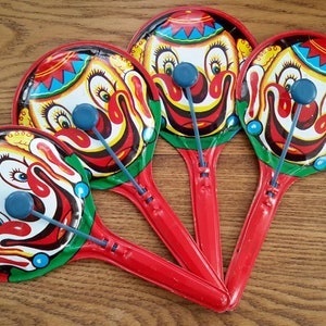 May include: Four red and white clown rattle toys with blue and green accents. Each rattle has a clown face with a big smile and a blue nose. The rattles are made of metal and have a handle.
