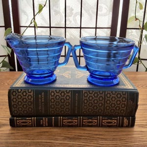 Peut inclure: Un pot à crème et un sucrier en verre bleu cobalt, avec des détails côtelés horizontaux et des poignées. Ils reposent sur une pile de livres anciens avec des lettres dorées. Le pot à crème a un bec verseur.