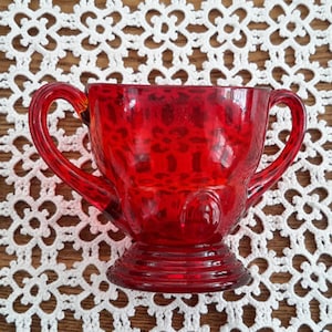 Peut inclure: Un bol à sucre en verre rouge avec deux anses. Le bol a une surface texturée et une base ronde.