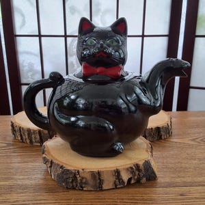 Peut inclure: Une théière en céramique noire brillante en forme de chat assis. Le chat présente des yeux verts, des oreilles intérieures rouges, un nœud papillon rouge et une anse en forme de queue courbée. Le bec verseur est sur le côté.