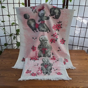 May include: A vintage pink and white towel set with poodle illustrations and red bows, plus floral accents. The set includes a large bath towel and a smaller hand towel, both with fringed edges.